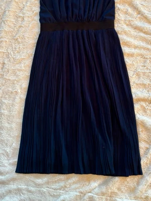 Marinblå plisserad klänning från Vila - Snygg marinblå klänning från Vila med plisserad kjol och markerad midja. Klänningen är ärmlös och har en rundad halsringning med knapp i nacken. Materialet känns lätt och har ett fint fall, perfekt för dig som gillar stilrena detaljer.