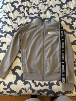 Grå Hoodrich ziptröja XL - Säljer en grå ziptröja från Hoodrich i storlek XL. Tröjan har lång ärm, ståkrage och dragkedja framtill. På vänster bröst finns Hoodrich-loggan i vitt och längs ena ärmen ett svart band med vit text. Materialet är 65% polyester och 35% bomull.