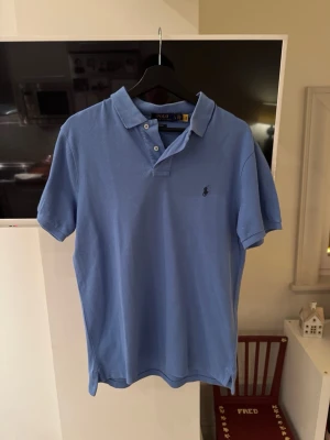 Blå pikétröja från Polo Ralph Lauren - Säljer en klassisk blå pikétröja från Polo Ralph Lauren i storlek L. Tröjan har korta ärmar, krage och två knappar framtill. Slim fit-modell. Tröjan är använd ett par gånger men har inga skador