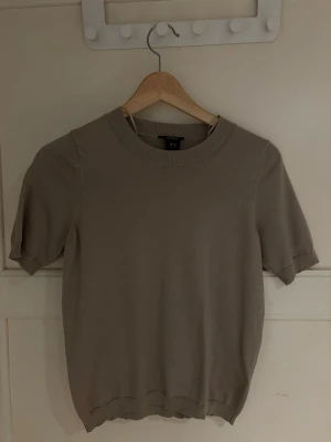 Brun/beige topp från Lindex storlek XS - Säljer en beige stickad topp från Lindex med rund hals och korta ärmar. Toppen har en enkel och stilren design med ribbade kanter vid hals, ärmslut och nederkant. Perfekt att matcha med jeans eller kjol!