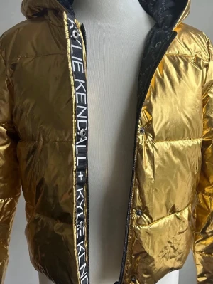 Guldig pufferjacka Kendall + Kylie XS - Säljer en glansig guldig pufferjacka från Kendall + Kylie i storlek XS. Jackan har huva, dragkedja och tryckknappar framtill samt ett coolt band med logotyp längs öppningen. Materialet är syntet och jackan är quiltad för extra värme.
