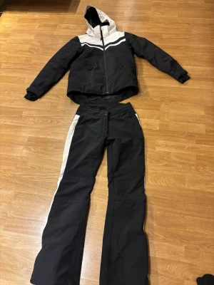 Vinterjacka + Svarta täckbyxor med vita ränder - Svart och vit vinterjacka + ett par svarta täckbyxor med vita sidopaneler. Byxorna har förstärkta benslut och praktiska fickor med dragkedja. Materialet är slitstarkt och vattenavvisande, perfekt för kalla dagar och vinteraktiviteter. Snygg och sportig look med raka ben. Jacka och byxa kan tvättas innan köp om önskas. Finns lite fläckar som förmodligen går bort i tvätt. Jacka stl 164 byxa stl 170, passar från 158. Finns lite ska. Från pjäxor, fler bilder tas efter önskemål.