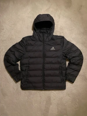 Svart pufferjacka från Adidas, M - Svart pufferjacka från Adidas med huva och dragkedja framtill. Jackan har Adidas-logga på bröstet och är tillverkad i polyester. Jätte bra för kalla vintrar. Inga defekter, pris kan diskuteras!