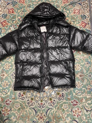 Svart pufferjacka från Moncler - Säljer en svart pufferjacka från Moncler med glansig finish och huva. Jackan har dragkedja framtill, två sidofickor med dragkedja och en ficka på ärmen med Moncler-logga. Perfekt för kalla dagar och riktigt snygg till streetwear.
