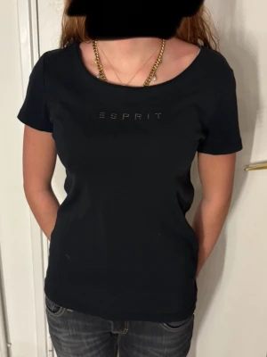 Svart t-shirt från Esprit - Snygg svart t-shirt från Esprit med korta ärmar och rund halsringning. Diskret logga framtill i små nitar som ger en cool touch. Perfekt till jeans eller kjol och enkel att styla med smycken. Gjord i mjuk bomull för skön känsla.
