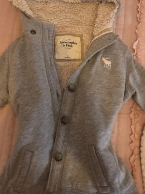 Grå hoodie från Abercrombie & Fitch - Mysig grå hoodie från Abercrombie & Fitch i storlek S. Tröjan har knappar framtill, huva med fluffigt foder och broderad logga på bröstet. På ena ärmen står Abercrombie i stora bokstäver. Perfekt för kyliga dagar och riktigt skön att ha på sig.