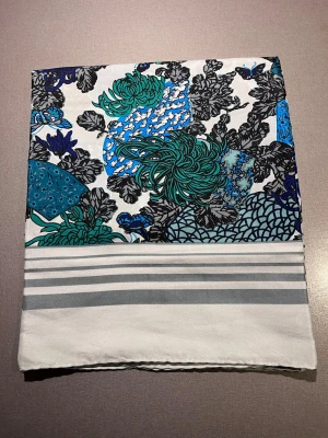 Blommig scarf  - Scarf med blommigt mönster i blått, grönt och svart på ljusblå botten. Gråa ränder längs nederkanten. Tillverkad i glansigt tyg som känns satin. Perfekt över tröjan, klänningen eller som detalj på väskan. Mått: ca88x66 cm. Mycket bra skick, som ny! Säljs på flera sidor, först till kvarn. 
