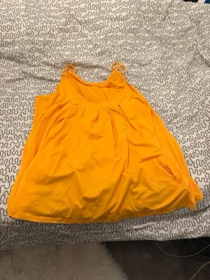 Gul ärmlös linne - Säljer en solgul linne med tunna axelband och rund halsringning. Linne är ärmlös och har en luftig, avslappnad passform med fina veck framtill. Perfekt för varma dagar och enkel att styla med accessoarer.
