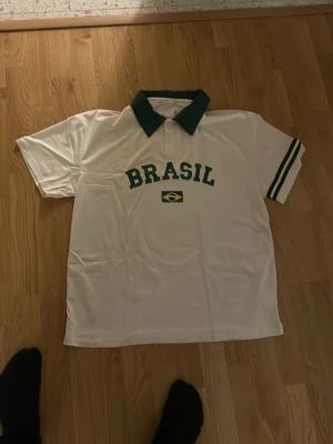 Vit Brasil pikétröja med grön krage - Snygg vit pikétröja med grön krage och gröna ränder på ärmarna. Tryck med texten 'BRASIL' och brasilianska flaggan på bröstet. Perfekt för dig som gillar fotboll eller vill ha en sportig stil.