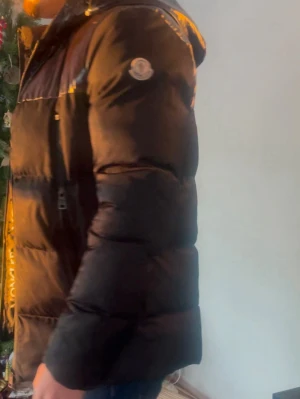 Svart Moncler pufferjacka med huva - Säljer en svart pufferjacka från Moncler med huva och stora logotexter längs dragkedjan. Jackan har dunfyllning, långärmade ärmar och en klassisk Moncler-logga på armen. Insidan av kragen är röd för en snygg kontrast. Perfekt för kalla dagar.