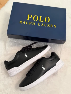 Svarta sneakers från Polo Ralph Lauren - Snygga svarta sneakers från Polo Ralph Lauren med vit sula och klassisk logga på sidan. Skorna har snörning, rund tå och är tillverkade i skinn med textilfoder och gummisula. Perfekta för dig som gillar stilrena och tidlösa sneakers.