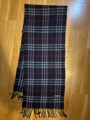 Burberry halsduk  - Snygg rutig scarf från Burberrys i mörkblått med grå och röda linjer. Den är tillverkad i 100% lammull och har klassiska fransar i nederkant. Perfekt nu i vinter ❄️ 
