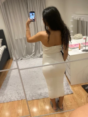 Vit axelbandslös ribbad långklänning - Säljer en superfin vit axelbandslös långklänning i ribbat material. Klänningen har en snygg detalj med twist framtill vid bysten och sitter figurnära. Perfekt för dig som gillar en stilren och enkel look med lite edge.