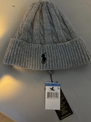 Grå kabelstickad mössa Polo Ralph Lauren - Snygg grå kabelstickad mössa från Polo Ralph Lauren med klassisk ribbad kant och broderad svart logga framtill. Mössan har en mjuk och varm känsla, perfekt för kyliga dagar. Taggarna sitter kvar och designen är tidlös.