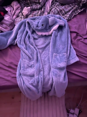 Blå  morgonrock  med hajhuva - Säljer en blå morgonrock med en rolig huva som ser ut som en haj, komplett med ögon och tänder. Västen har två stora fickor framtill och är supermjuk och mysig. Perfekt för chill dagar hemma eller när du vill sticka ut lite extra.