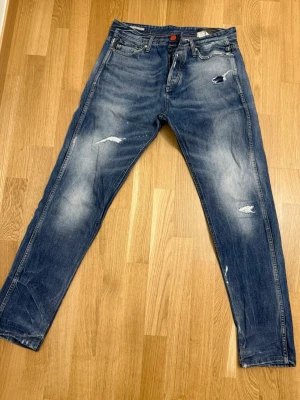 Blå jeans från Jack & Jones - Säljer ett par blå jeans från Jack & Jones, modell Erik Original Felling. Jeansen har slitna detaljer och snygga tvättade partier på både fram- och baksida. Klassisk femficksdesign och orange knapp framtill. Perfekt för dig som gillar en avslappnad streetstil.