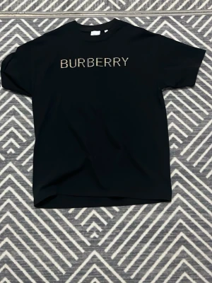 Svart Burberry t-shirt med logga - Svart t-shirt från Burberry med klassisk rund hals och korta ärmar. Stor Burberry-logga framtill i rutigt mönster som ger en snygg kontrast mot den svarta bakgrunden. Materialet är mjukt och känns skönt mot huden, perfekt för dig som gillar stilrena designer med en twist.
