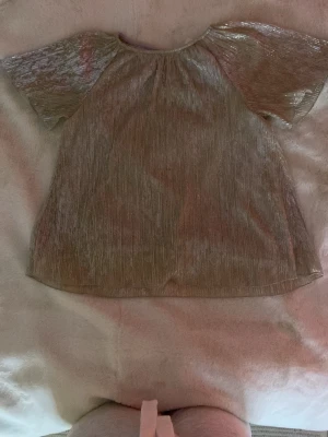Glittrig guld blus med kort ärm - Säljer en glittrig guldblus med korta, vida ärmar och rund hals. Tyget har en skimrande effekt och känns lätt och luftigt, perfekt för dig som vill sticka ut lite extra. Passar till både jeans och kjol för en trendig look.