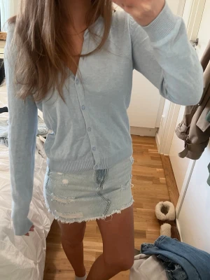 Ljusblå kofta med knappar - Supersöt ljusblå kofta med v-ringning och knappar hela vägen framtill. Långärmad modell i mjukt, tunt material som passar perfekt till både jeans och kjol. Ribbstickade muddar vid ärmslut och nederkant för en clean look.