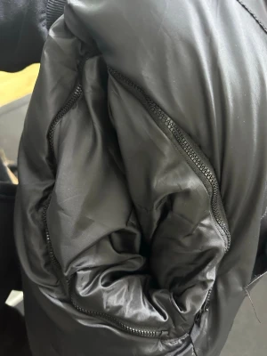 Svart pufferjacka från Burberry - Säljer en svart pufferjacka från Burberry med huva och klassiskt rutigt foder. Jackan har två dragkedjefickor på framsidan och en snygg Burberry London England patch på ärmen. Perfekt för kalla dagar och har en stilren look.