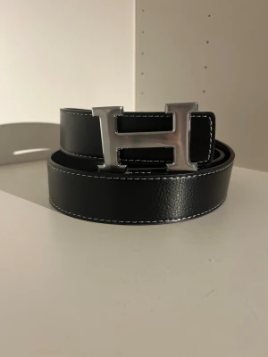 Svart skärp med H-spänne i skinn - Snyggt svart skärp i skinn med ett stort, silvrigt H-format metallspänne. Skärpet har vit kontrastsöm längs kanterna och en stilren, klassisk look. Perfekt för att lyfta din outfit med en exklusiv touch.