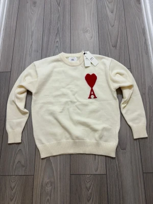 Vit stickad tröja från Ami - Säljer en vit stickad tröja från Ami Alexandre Mattiussi i storlek M. Tröjan har ett rött hjärta med bokstaven A broderat på bröstet, rund hals och ribbade muddar vid ärmslut och nederkant. Materialet är mjuk bomull och passformen är normal.