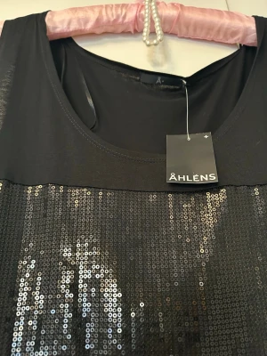 Svart paljettlinnetopp från Åhléns - Snygg svart ärmlös linnetopp från Åhléns med glittriga paljetter framtill. Toppen har rund halsringning och är tillverkad i mjuk viskos. Perfekt för att sticka ut med lite extra bling! Paljetterna ger en cool och festlig vibe.