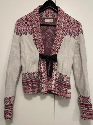 Odd Molly kofta med mönster och rosett - Superfin kofta från Odd Molly i ljusgrått med rosa och vinröda detaljer. Koftan har ett broderat mönster över axlar, ärmar och nederkant samt en svart rosett framtill. Långärmad och stickad i mjukt material, perfekt för höst och vinter.