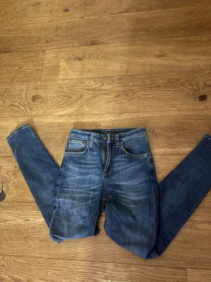 Blå jeans från Nudie Jeans - Säljer ett par klassiska blå jeans från Nudie Jeans med ikoniska bakfickor med vågigt mönster. Jeansen har normal passform, raka ben och perfekta till vardagslooken!