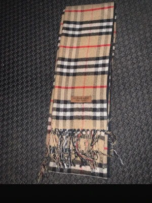 Rutig beige halsduk från Burberry - Klassisk halsduk från Burberry i beige, svart, vit och röd med ikoniskt rutmönster. Halsduken har fransar i ändarna och en snygg Burberry-label. Perfekt accessoar för att lyfta din outfit.