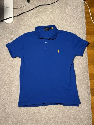 Blå pikétröja från Polo Ralph Lauren - Klassisk blå pikétröja från Polo Ralph Lauren i slim fit. Tröjan har korta ärmar, krage och två knappar framtill. Ikoniska gula loggan broderad på bröstet. Perfekt till jeans eller shorts för en clean och stilren look.