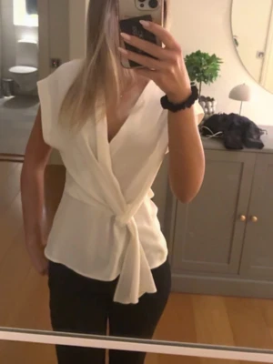 Vit omlottopp med knyt - Säljer en stilren vit omlottopp med knyt framtill. Toppen har v-ringning, ärmlös design och ett mjukt, lätt tyg som ger en snygg siluett. Perfekt till jeans eller kjol för en fräsch look.