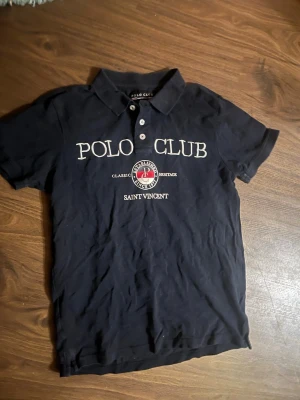 Marinblå Polo Club t-shirt S - Snygg marinblå t-shirt från Polo Club Saint Vincent med klassisk krage och knappar framtill. Stor logga och text på bröstet i vitt samt ett broderat emblem i rött och vitt. Perfekt till en avslappnad men ändå stilren look.