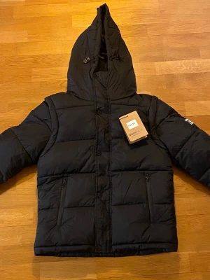 Svart pufferjacka från Burberry - Säljer en svart pufferjacka från Burberry med huva och dragkedja. Jackan har två fickor med dragkedja, Burberry-logga på ärmen och klassiskt rutigt foder i huvan. Tillverkad i polyester och har en riktigt skön, vadderad känsla. Perfekt för kalla dagar.