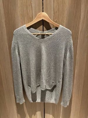 Grå stickad tröja från Lindex - Säljer en grå stickad tröja från Lindex i storlek M. Tröjan har v-ringning framtill och snygg detalj med korsade band i nacken. Långärmad modell med ribbade muddar och avslappnad passform. Perfekt till höst och vinter!