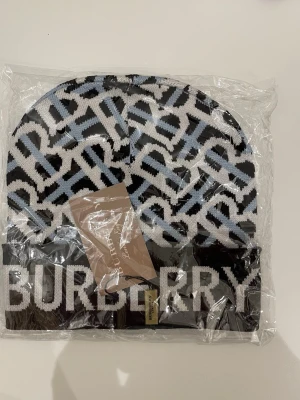 Burberry mönstrad stickad mössa - Snygg stickad mössa från Burberry med ett grafiskt mönster i svart, vitt och ljusblått. Mössan har bred ribbad kant nedtill och stor Burberry-logga framtill. Perfekt för dig som vill ha en statement piece i garderoben.