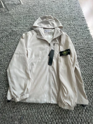 Beige vindjacka från Stone Island - Säljer en beige vindjacka från Stone Island med huva och dragkedja framtill. Jackan har den klassiska Stone Island-loggan på vänster ärm och elastiska muddar vid ärmslut och nederkant. Tillverkad i lätt polyester, perfekt för lager-på-lager och streetwear.