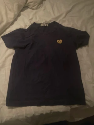 Svart t-shirt Comme des Garçons Play - Svart t-shirt från Comme des Garçons Play med det ikoniska hjärte-logotypen i gult på bröstet. Klassisk passform och rund hals. Tillverkad i mjuk bomull som känns skön mot huden. Perfekt för dig som gillar stilrena plagg med en twist.