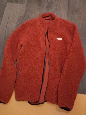 Rostfärgad teddyjacka True North M - Mysig teddyjacka från True North i en snygg rostbrun färg. Jackan har dragkedja framtill, två fickor och en mjuk, fluffig yta som håller dig varm. Perfekt för kyliga dagar och enkel att matcha med din vardagsstil.