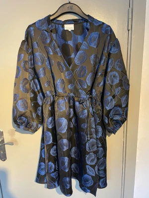 Blommig omlottklänning från Noella - Supersnygg kort omlottklänning från Noella med blått blommönster på svart botten. Klänningen har puffiga långärmar, v-ringning och knyt i midjan för en skön passform. Materialet känns glansigt och lyxigt, perfekt för dig som vill sticka ut.