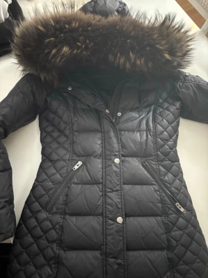 Rock and blue jacka med päls  - Lång svart pufferjacka med quiltat mönster och stor huva med fluffig päls. Jackan har ett hål i fodret men kan lätt lagas och påverkar inte jackans funktion och syns inte. 
