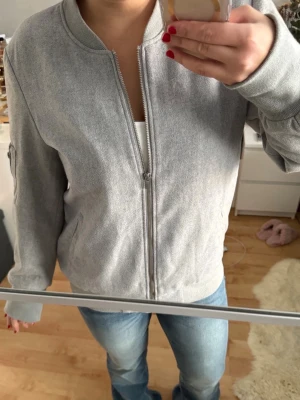 Grå bomberjacka med dragkedja - Snygg grå bomberjacka med klassisk rund krage och silverfärgad dragkedja framtill. Jackan har långärmade ärmar, ribbade muddar och fickor på sidorna. Perfekt för dig som gillar en skön och mjuk jacka med en enkel och stilren look. Den är i strl L men passar bra för de flesta storlekar, bär själv S. 
