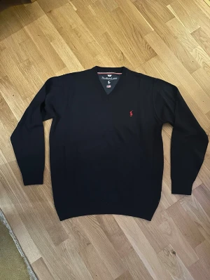 Svart v-ringad tröja Polo Ralph Lauren - Klassisk svart v-ringad tröja från Polo Ralph Lauren med röd broderad logga på bröstet. Tröjan har långa ärmar och ribbade muddar vid ärmslut och nederkant. Perfekt till lager på lager och enkel att matcha med allt.