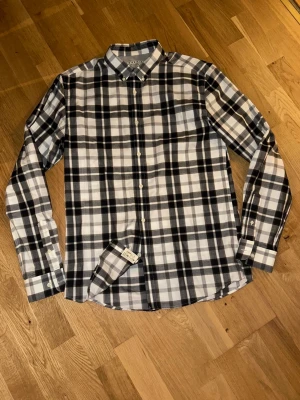 Rutig skjorta från Jack & Jones - Säljer en snygg rutig skjorta från Jack & Jones Premium i svart, vit och grå. Skjortan har klassisk krage, långa ärmar och knappar framtill. Perfekt för dig som gillar stilrena och tidlösa plagg.