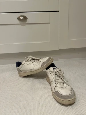 Vita sneakers från Axel Arigato - Säljer ett par stilrena vita sneakers från Axel Arigato med gråa detaljer och mörkblå insida. Skorna har klassisk snörning, perforerade sidor och en platt sula. Tillverkade i skinn och mocka för en clean och modern look. Inga defekter på skorna och de är endast använda ett fåtal gånger. Pris kan diskuteras. 