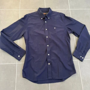 Mörkblå skjorta från Morris L - Klassisk mörkblå skjorta från Morris med button down-krage och vita knappar. Broderad logga på bröstet och lång ärm. Skjortan är i bomull och har en stilren, tidlös look som funkar till många olika outfits.