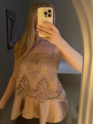 Champagnefärgad spetslinne i satin - Supersnygg champagnefärgad linnetopp i satin med tunna axelband och broderade spetsdetaljer framtill. Toppen har en lätt utsvängd nederdel och romantisk känsla, perfekt att styla med jeans eller kjol. 💛