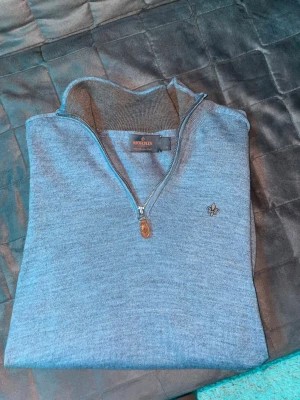 Grå half zip tröja från Morris - Säljer en stilren grå tröja från Morris med half zip och liten broderad logga på bröstet. Tröjan är stickad och har en hög krage som ger en clean look. Perfekt för dig som gillar klassisk men modern stil.