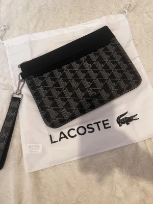 Svart kuvertväska från Lacoste - Snygg svart kuvertväska från Lacoste med geometriskt mönster i grått och diskret logga framtill. Väskan har avtagbar handledsrem och dragkedja upptill. Tillverkad i tåligt syntetmaterial och perfekt för att bära det viktigaste. Helt ny och dustbag medföljer.