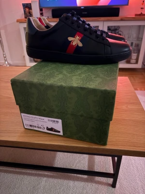 Gucci Ace sneakers med bi-detalj - Säljer ett par svarta Gucci Ace sneakers i skinn med klassiska röda och blå ränder på sidan och broderad guldfärgad bi. Baksidan har metallicröd och metallicblå hälflik med ormmönster och Gucci-logga. Skorna har rund tå och svarta snören.